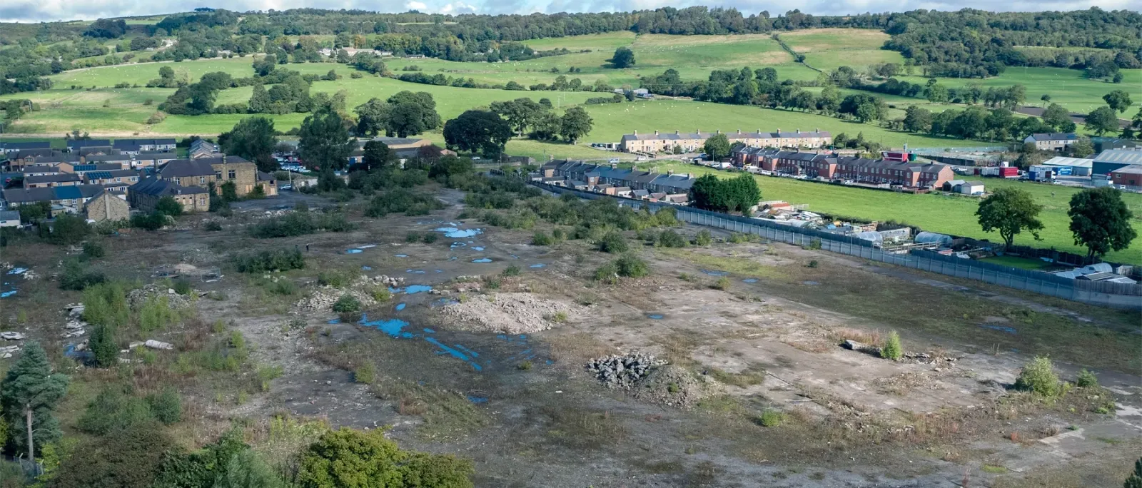 An aerial view of Wolsingham steel works, a large, vacant lot with patches of rubble, puddles, and sparse vegetation. In the background, rows of houses, industrial buildings, and green fields are visible. The area is surrounded by rolling hills, trees, and countryside, with a fenced boundary separating the lot from nearby structures. The sky is partly cloudy with sunlight illuminating the landscape.